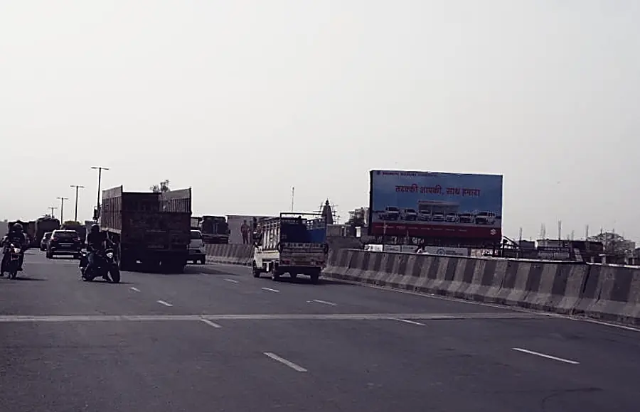 A highway hoarding beside an Agra arterial road, traffic on the carriageway under open sky